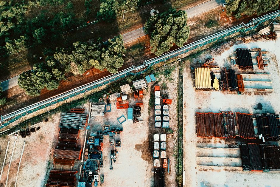 Visão aérea de um depósito de materiais de construção ao lado de uma área com vegetação e estrada, destacando a logística e armazenamento de produtos.