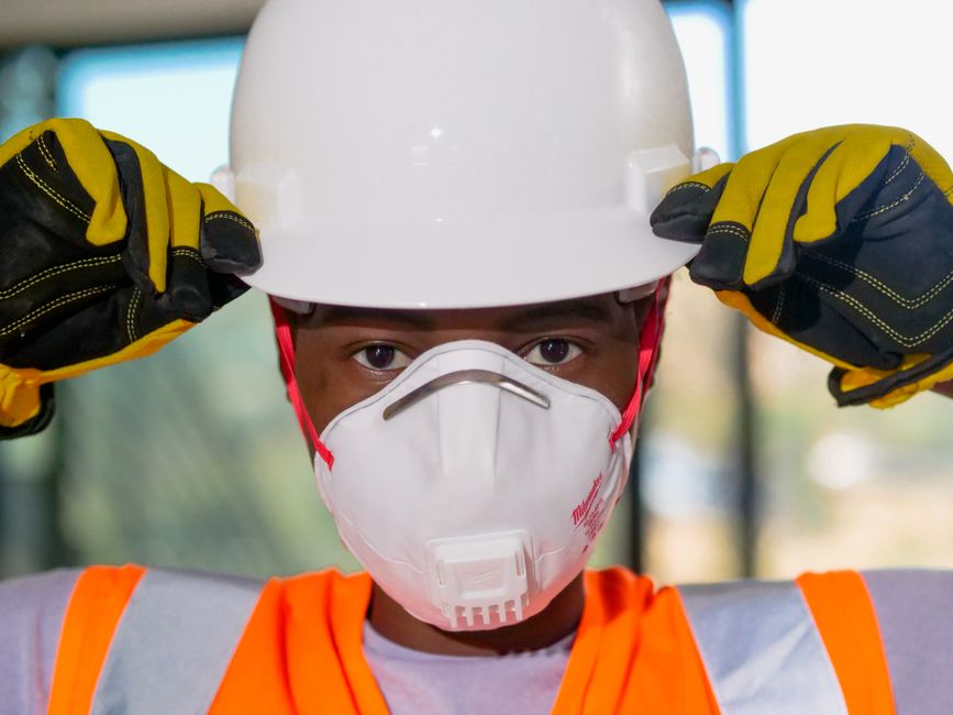 Imagem de um trabalhador da construção civil usando equipamentos de proteção individual, incluindo capacete, luvas e máscara, em ambiente de obra ou setor industrial.