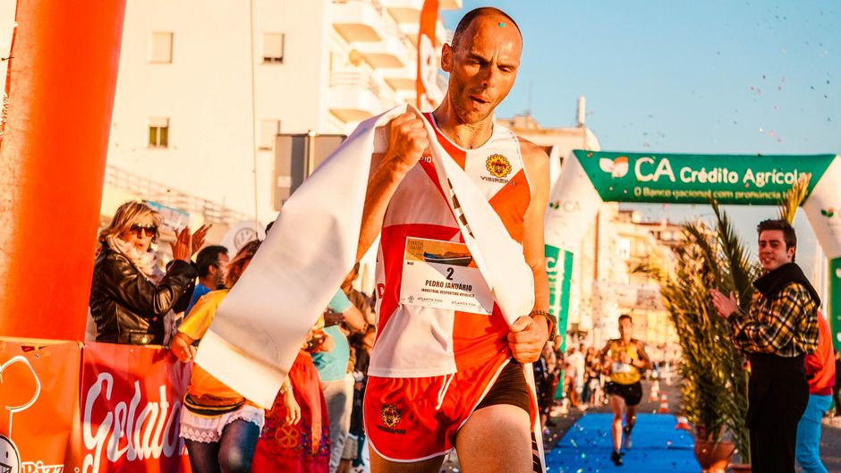 Atleta de corrida comemorando vitória em evento ao ar livre com público admirado ao fundo.