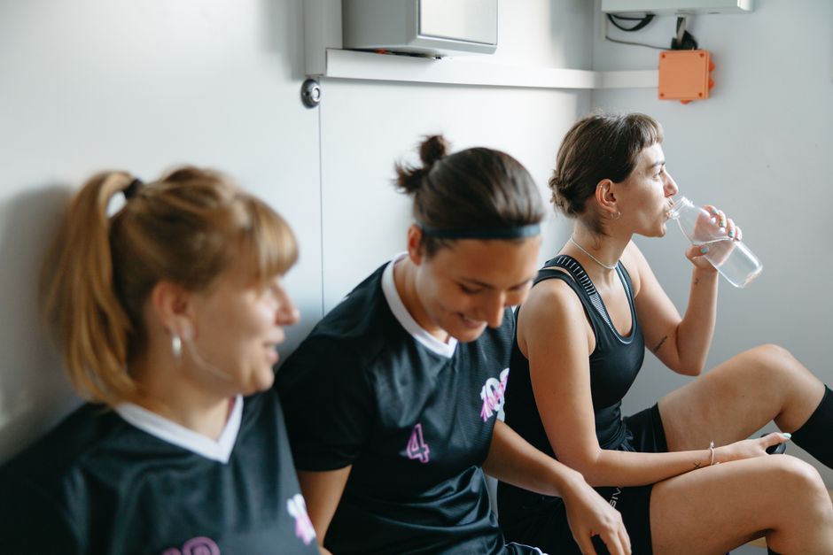 Três jogadoras de voleibol descansando após uma partida, sentadas contra a parede, uma delas bebendo água, demonstrando cansaço e alívio.