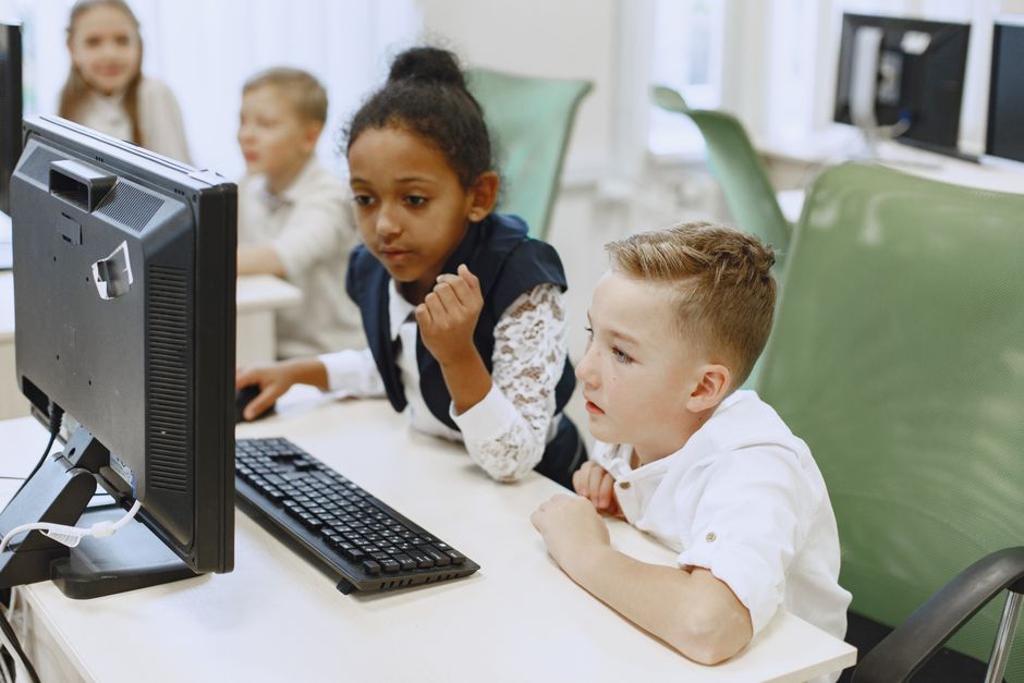 Grupo de crianças estudando em computador em uma sala de aula moderna, ilustrando aprendizagem digital e inclusão tecnológica.