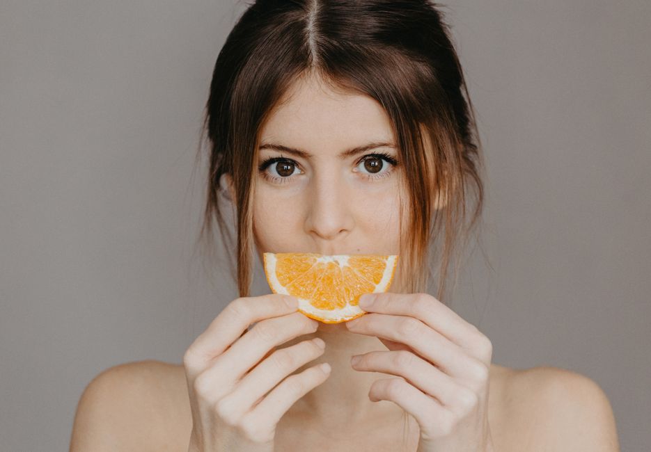 Imagem de uma mulher segurando uma fatia de laranja na boca, transmitindo uma sensação de frescor e saúde, ideal para artigos sobre alimentação saudável e bem-estar.