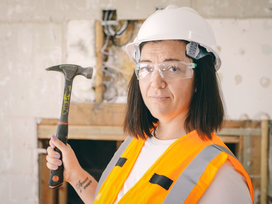 Mulher engenheira em ambiente de construção, usando capacete e colete de segurança, segurando um martelo e demonstrando determinação profissional.