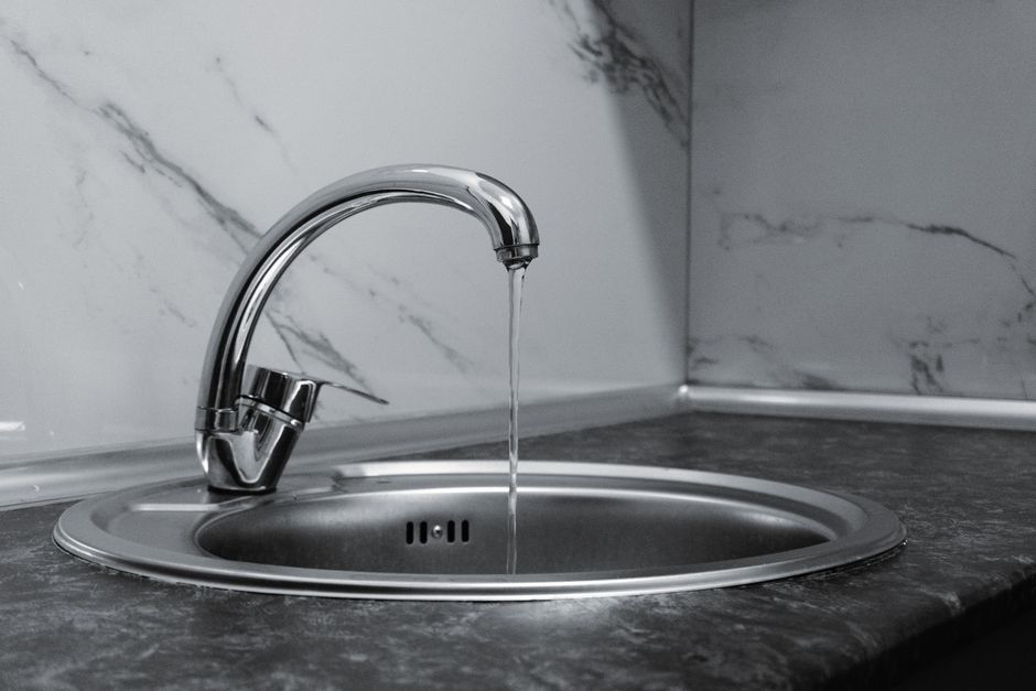 Imagem de uma pia de cozinha com torneira de metal por onde escorre água, sobre bancada de granito com revestimento de mármore.