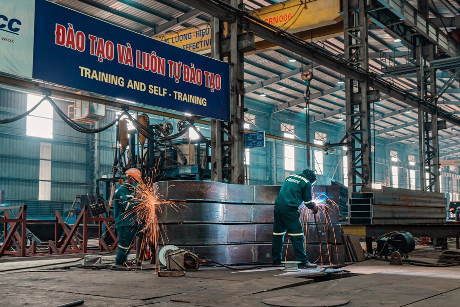 Imagem de uma fábrica industrial com operários realizando soldagem e montagem de grandes peças metálicas, dentro de um ambiente de produção e manufatura.
