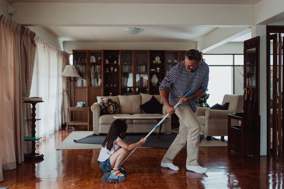 Pai e filha brincando de luta usando vassoura em sala de estar, transmitindo diversão familiar e conexão emocional.
