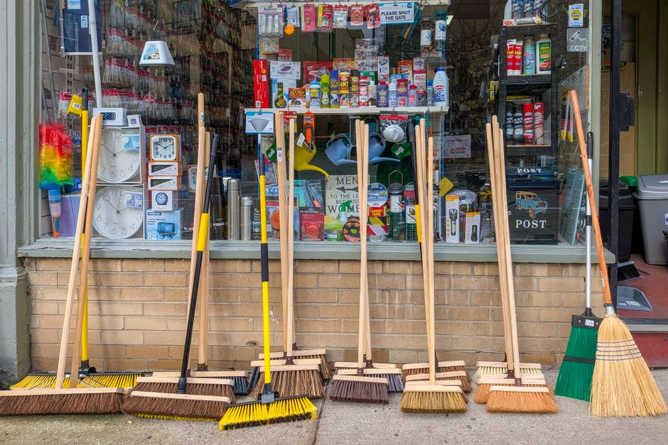 Varejo de limpeza e utensílios domésticos exibindo vassouras, espanadores e outros itens na fachada de uma loja.