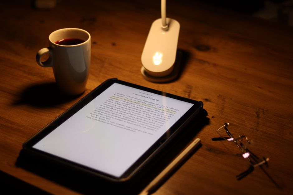 Imagem de um ambiente de leitura ou estudo noturno, com tablet, luz de leitura, café e óculos sobre uma mesa de madeira, criando um clima de concentração e trabalho remoto.