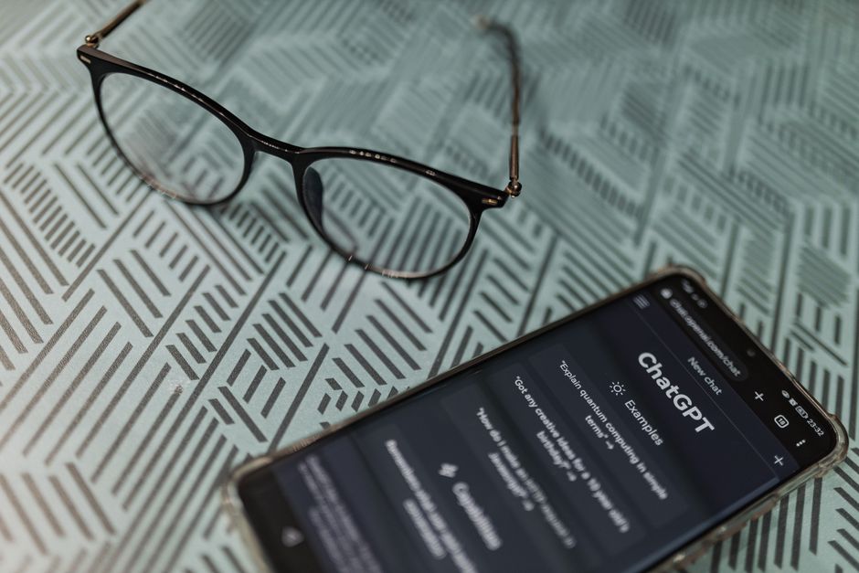 Imagem de um par de óculos e um celular com a tela do ChatGPT, sobre uma superfície com texto impresso, sugerindo pesquisa e uso de tecnologia para aprendizagem ou trabalho intelectual.