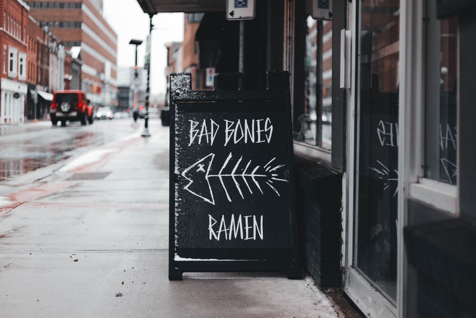 Placa de rua com anúncio de comida japonesa, mencionando 'Bad Bones Ramen', numa rua urbana durante o dia.