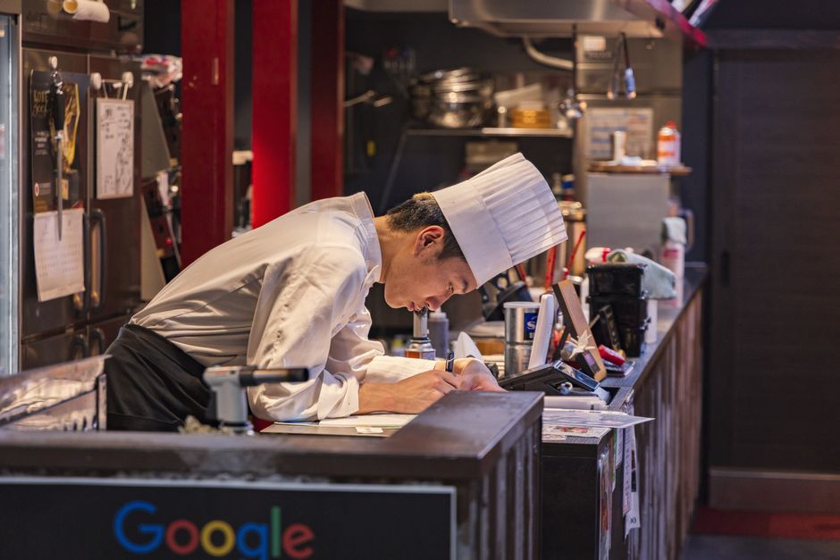 Chef focado anotando pedidos ou planejando tarefas em uma cozinha profissional, destacando o ambiente gastronômico e a dedicação no trabalho culinário.