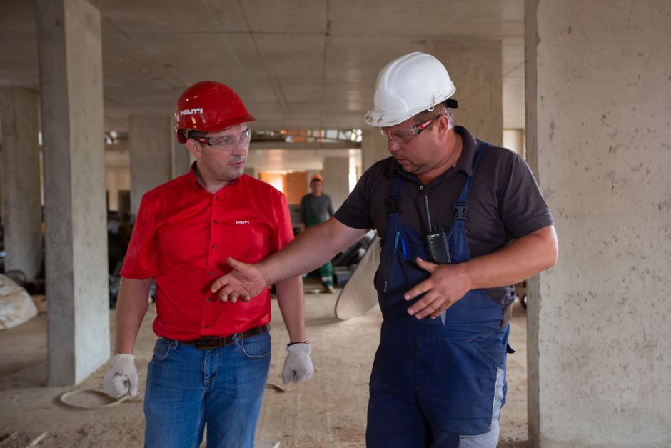 Homem explicando detalhes de uma obra em construção a outro profissional, em ambiente de obra civil em andamento.