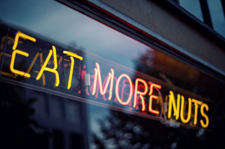 Letreiro de neon com a frase 'Eat More Nuts' em um ambiente urbano, transmitindo uma mensagem de incentivo a um estilo de vida saudável e alimentar-se de nozes.