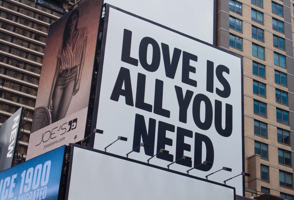 Placa com a mensagem inspiradora 'Love is all you need' em um cenário urbano de prédios altos, transmitindo uma ideia de amor e positividade no ambiente da cidade.