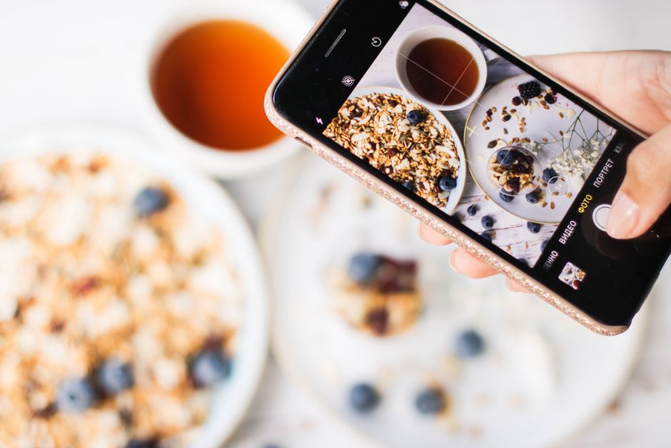Pessoa tirando foto de um café da manhã com granola, frutas e bebida, em um ambiente claro e acolhedor.