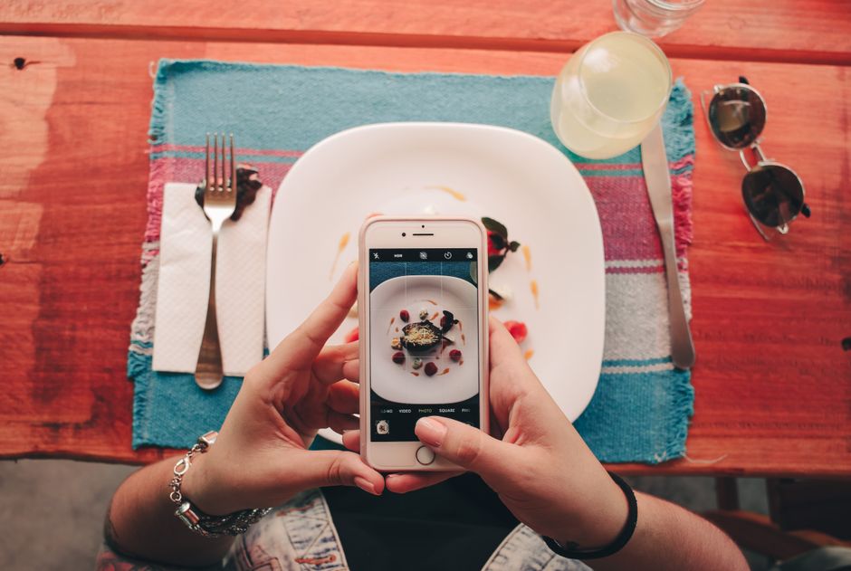 Pessoa tirando uma foto de um prato de sobremesa com frutas e sobremesa decorada, em um ambiente de restaurante ou café.