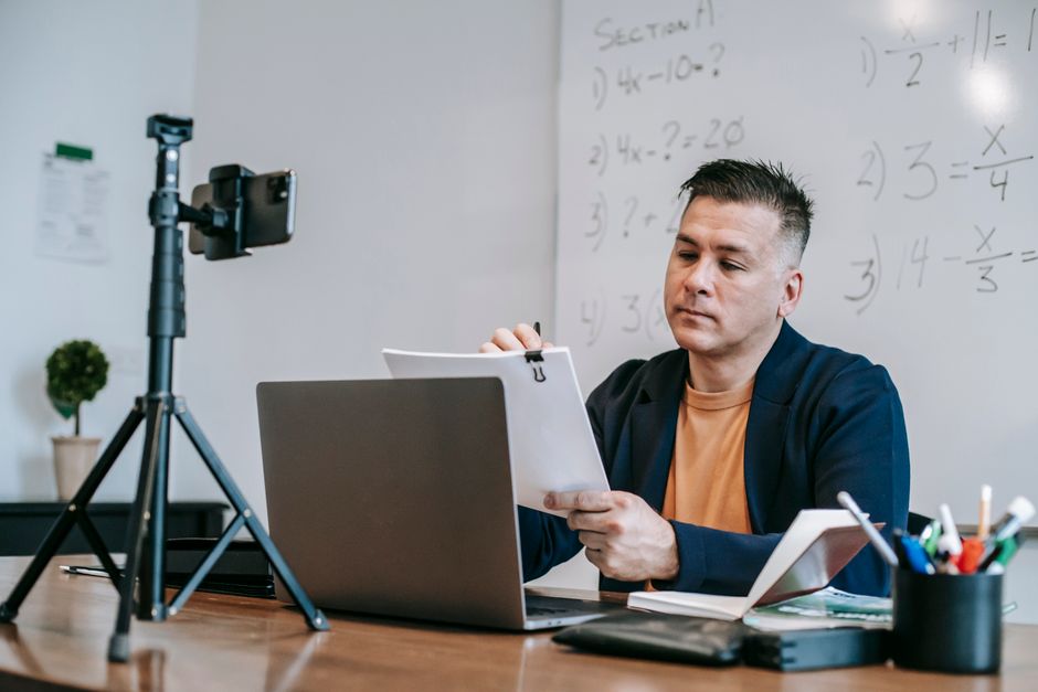 Professor ou professoras gravando aula online em sala de aula, com quadro de matemática ao fundo.