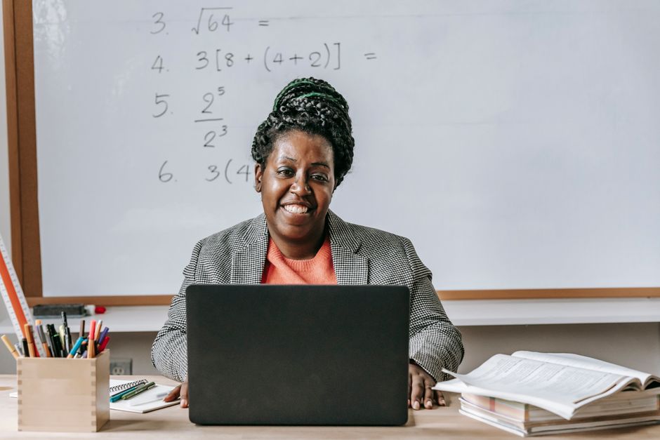 Professor de matemática em sala de aula, usando computador e ajudando alunos com exercícios de matemática.