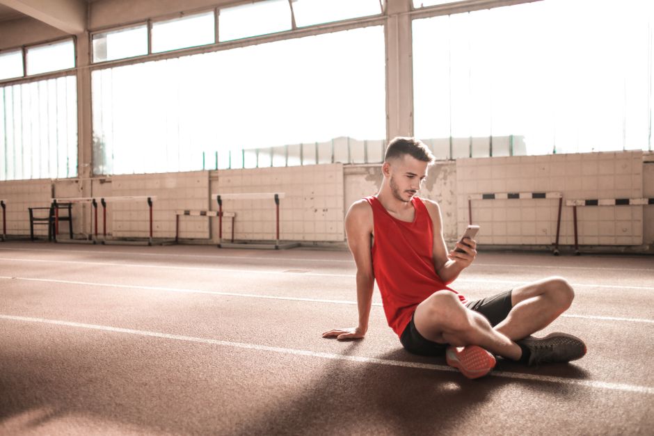 Jovem atleta sentado no piso de uma pista de corrida indoor, usando roupas esportivas e observando seu celular, em um ambiente de treinamento ou competição.