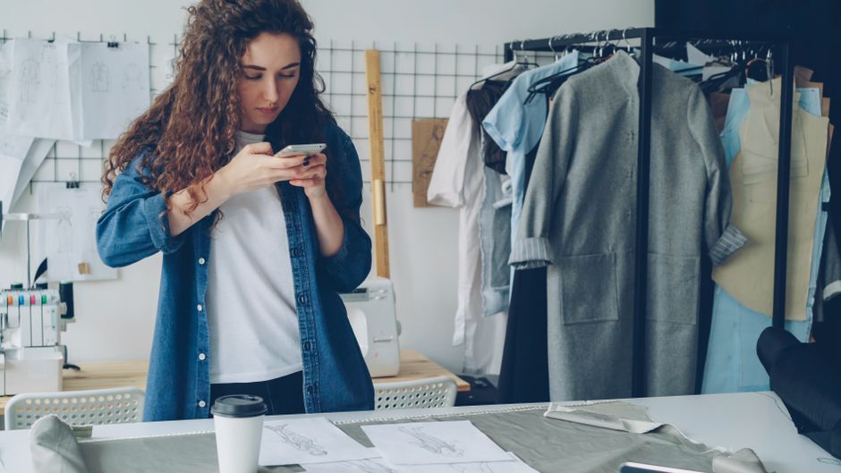 Jovem profissional visualizando uma mensagem no celular em um ambiente de criação de moda ou confecção, com roupas e desenhos ao fundo.
