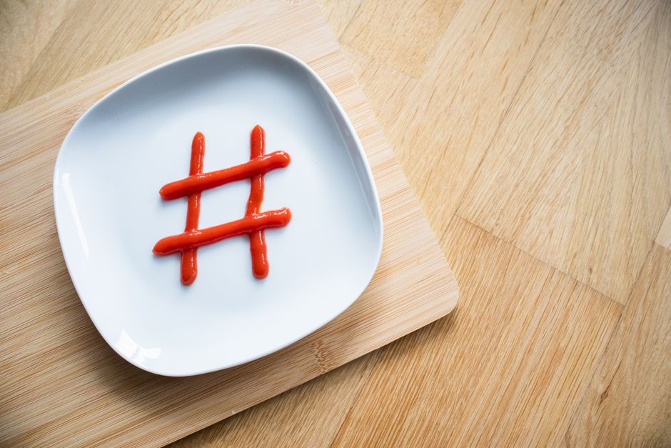 Imagem de um prato branco com um símbolo de hashtag feito com ketchup, sobre uma tábua de madeira, sugerindo tendências de redes sociais ou marketing digital.