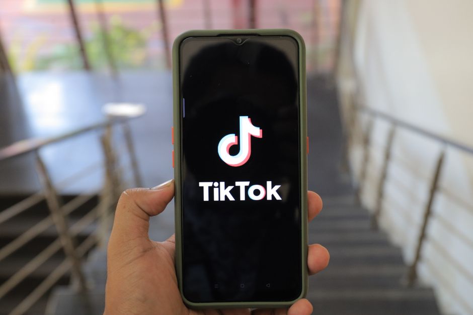 Pessoa segurando um smartphone exibindo o logo do TikTok, em ambiente com escadas e iluminação natural ao fundo.