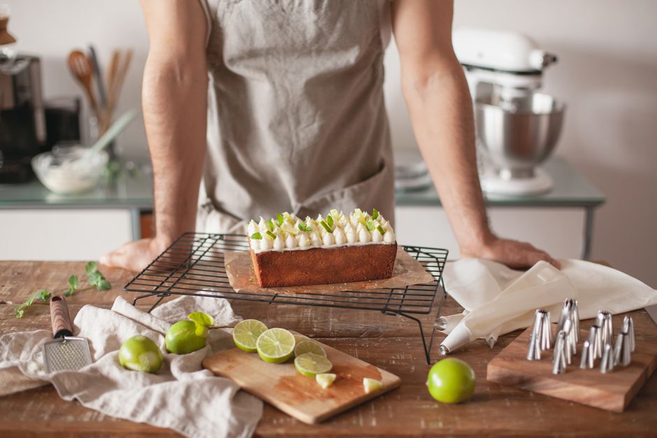 Homem preparando um bolo decorado com creme e ervas, em uma cozinha com utensílios de confeitaria ao fundo.