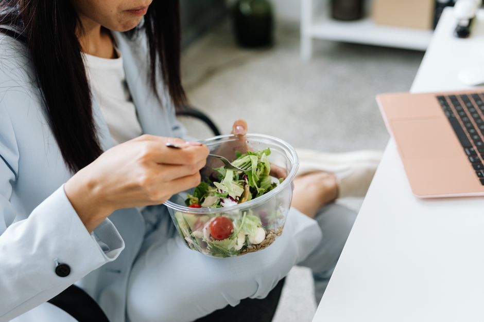 Pessoa trabalhando no escritório enquanto faz uma pausa para comer uma salada, simbolizando equilíbrio entre saúde e trabalho.