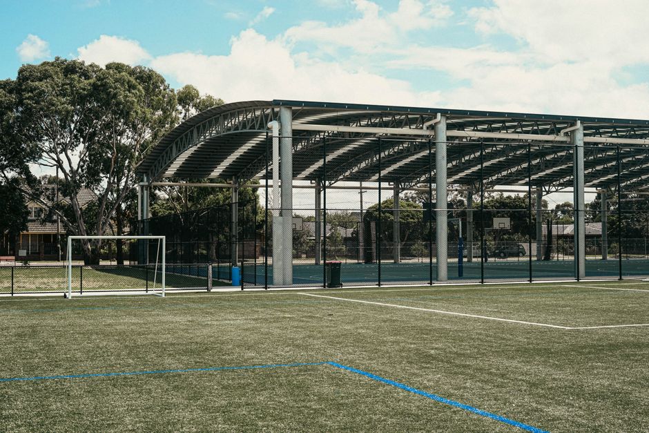 Imagem de uma quadra coberta ao ar livre, destinada a esportes, com destaque para a estrutura metálica do telhado e o campo de gramado.