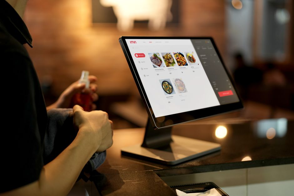Pessoa fazendo pedido em um restaurante usando um computador de mesa com um site de pedidos de comida, ilustrando tecnologia no setor de alimentação.