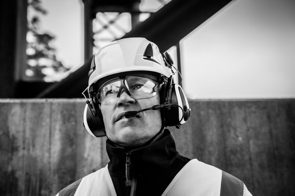 Homem com equipamento de segurança, incluindo capacete e headset, em ambiente urbano, possivelmente trabalhador da construção civil ou engenheiro no local de obra.