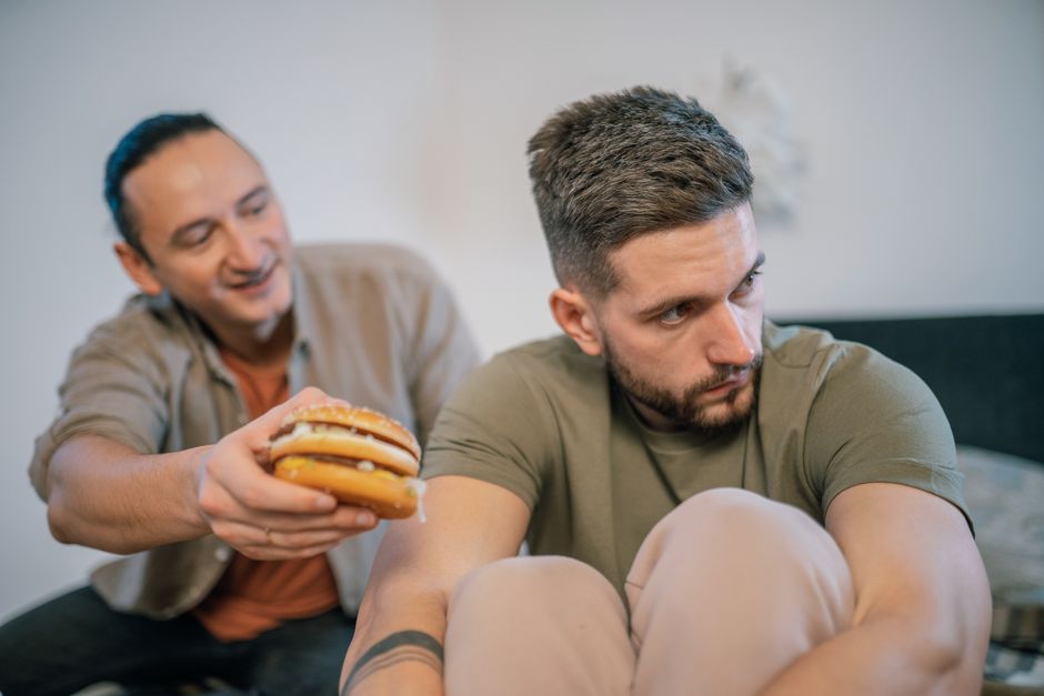Homem pensativo e desconfortável enquanto outro tenta aliviar sua tristeza com um sanduíche, ilustrando o tema de apoio emocional e dificuldades pessoais entre colegas.