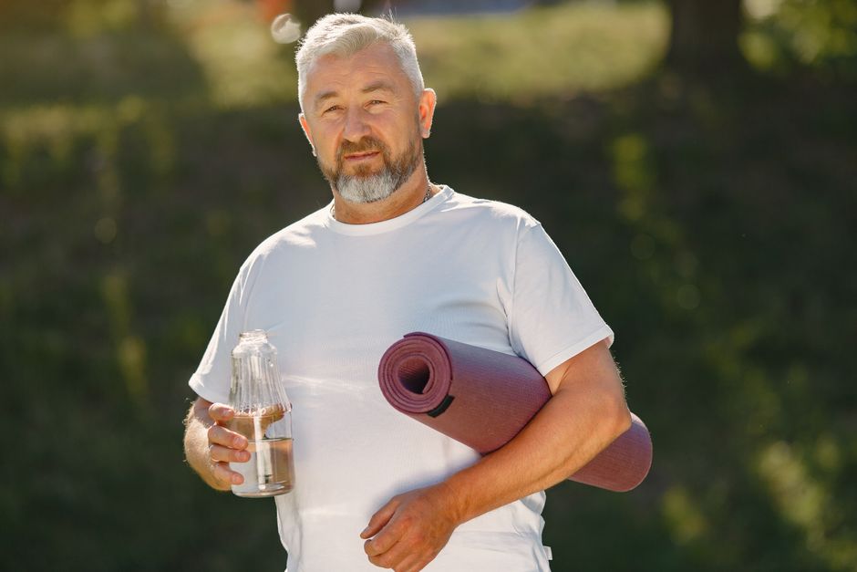 Homem idoso com aparência saudável segurando uma garrafa de água e um tapete de yoga, em ambiente ao ar livre, promovendo estilo de vida ativo e bem-estar.