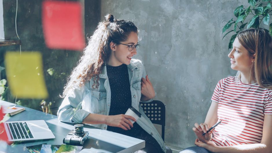 Duas mulheres conversando em um ambiente de trabalho colaborativo, com elementos que sugerem criatividade e reunião de equipe.