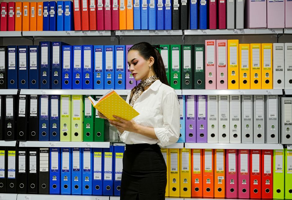Mulher analisando documentos em prateleira de arquivos coloridos, representando organização e gestão de documentos no ambiente de escritório.