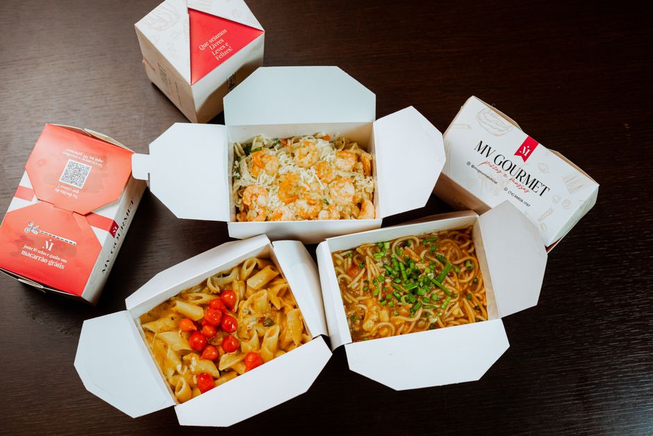 Imagem de três embalagens de comida para viagem contendo pratos de macarrão, salada e molho, sobre uma mesa de madeira, ideal para ilustrar temas de alimentação, delivery ou conveniência rápida.