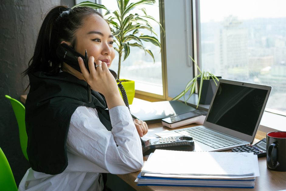 Mulher em ambiente de trabalho urbano, usando telefone e computador, representando comunicação e produtividade corporativa.