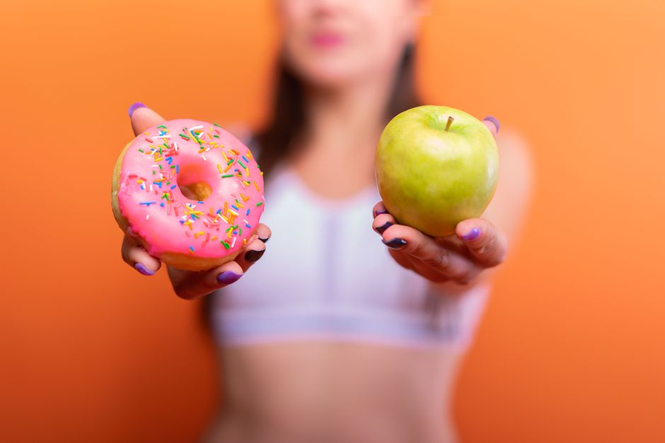 Imagem de uma mulher segurando um donut com cobertura rosa e confeitos coloridos em uma mão, e uma maçã verde na outra, contra um fundo laranja vibrante.