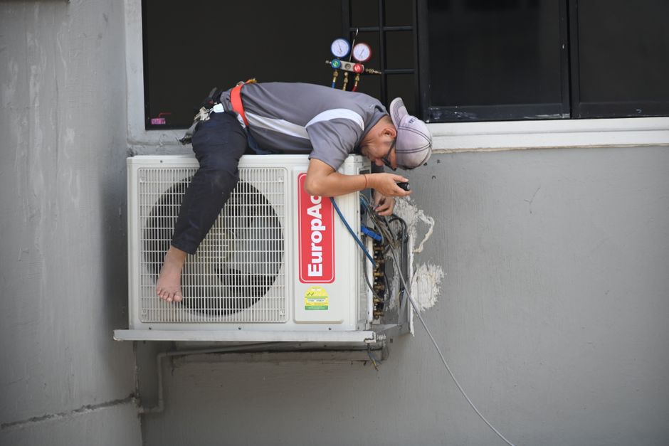Homem realizando manutenção em unidade de ar-condicionado instalado no exterior de um edifício, destacando a importância da manutenção preventiva em sistemas de climatização corporativa.