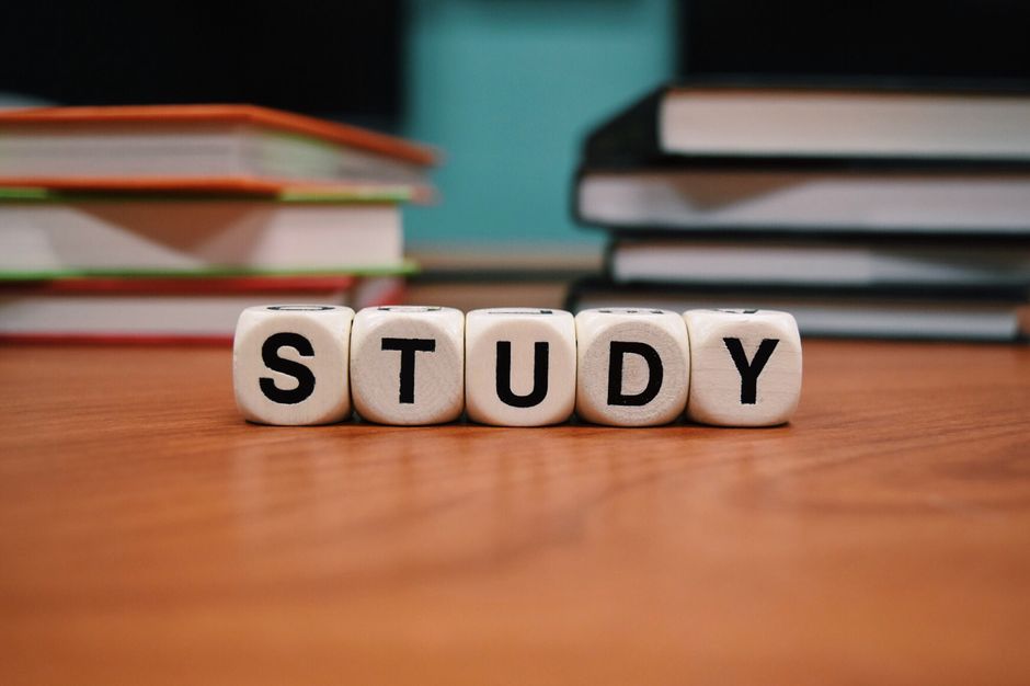 Imagem que representa o tema de estudo e aprendizagem, com blocos de letras formando a palavra 'STUDY' sobre uma mesa com pilhas de livros ao fundo.