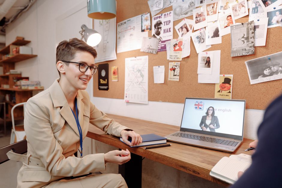 Mulher participando de aula de inglês online em ambiente de escritório ou espaço de estudo, com quadro de avisos na parede ao fundo.