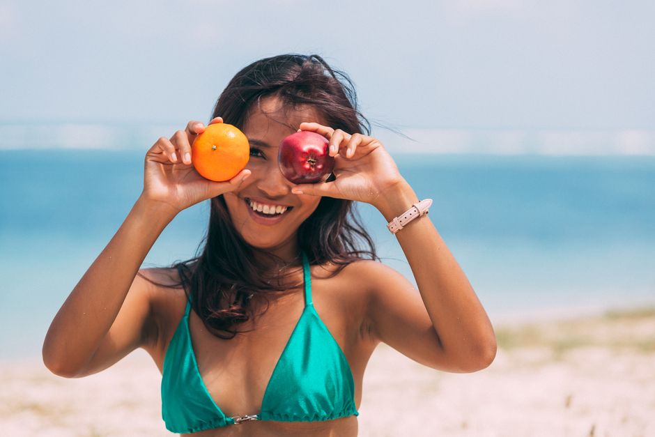 Mulher sorridente na praia segurando uma maçã e uma laranja na frente do rosto, transmitindo alegria e descontração ao ar livre.