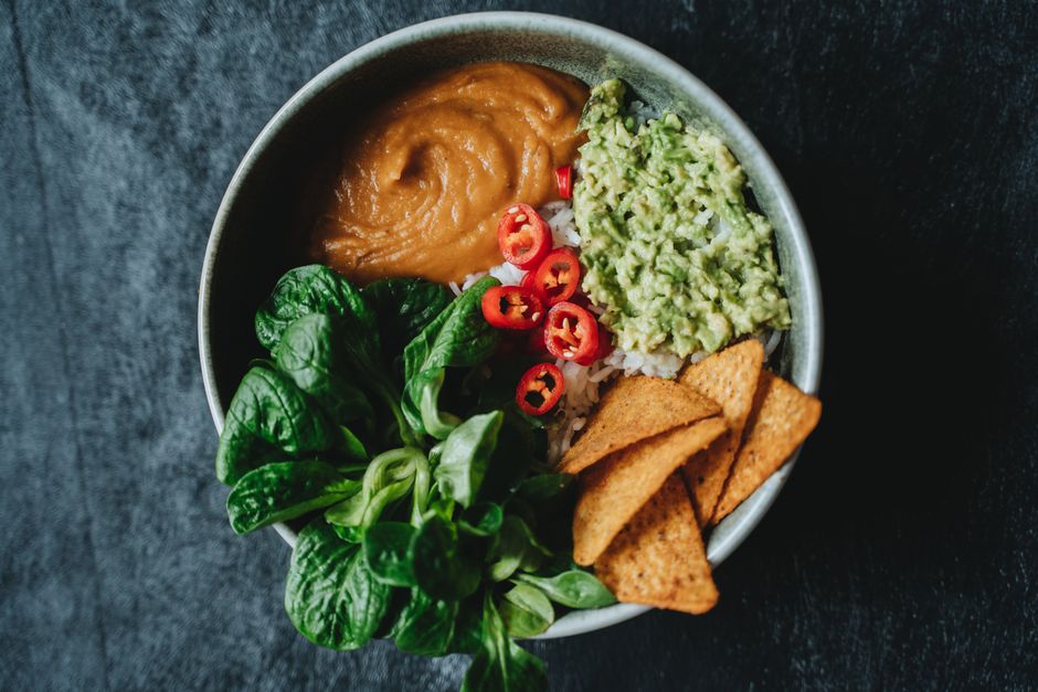 Imagem de uma tigela com comida saudável contendo guacamole, molho de feijão, alface, rodelas de pimenta e tortillas, com fundo escuro.