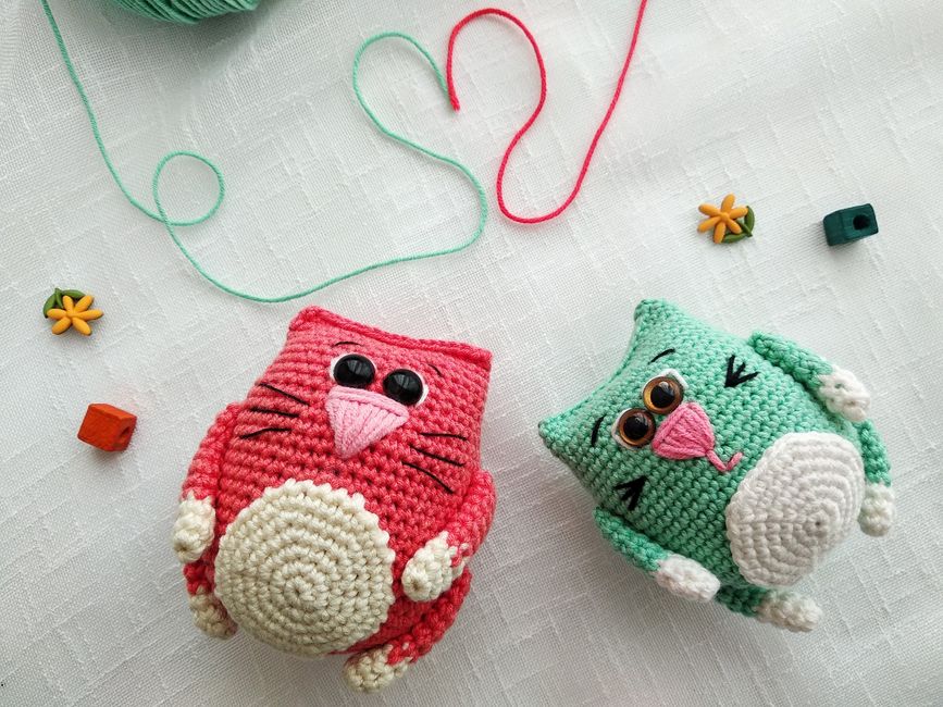 Imagem de dois brinquedos de amigurumi em formato de gatos fofos, com detalhes de olhos e bochechas, sobre uma superfície de tecido claro.