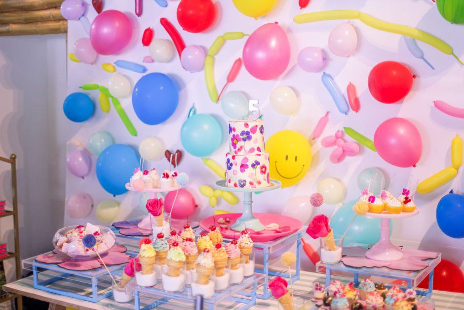 Cena de festa infantil decorada com balões coloridos, bolo central decorado e várias doces atraentes em um ambiente vibrante e alegre.