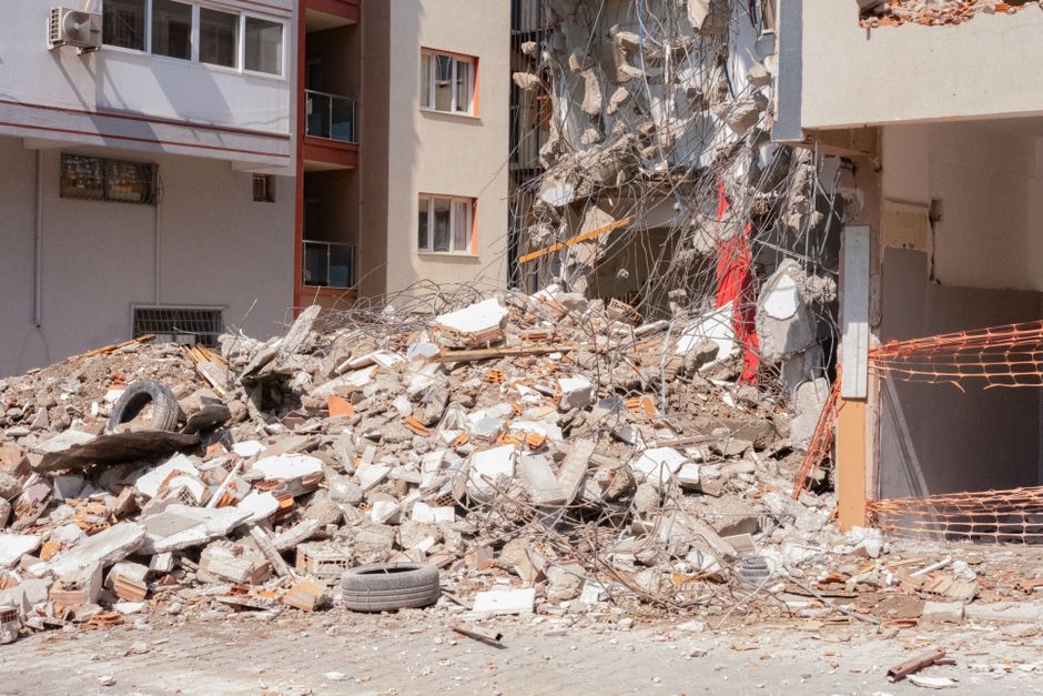Imagem de um prédio parcialmente destruído, com destroços e escombros ao redor, evidenciando os efeitos de uma calamidade ou desastre urbano.