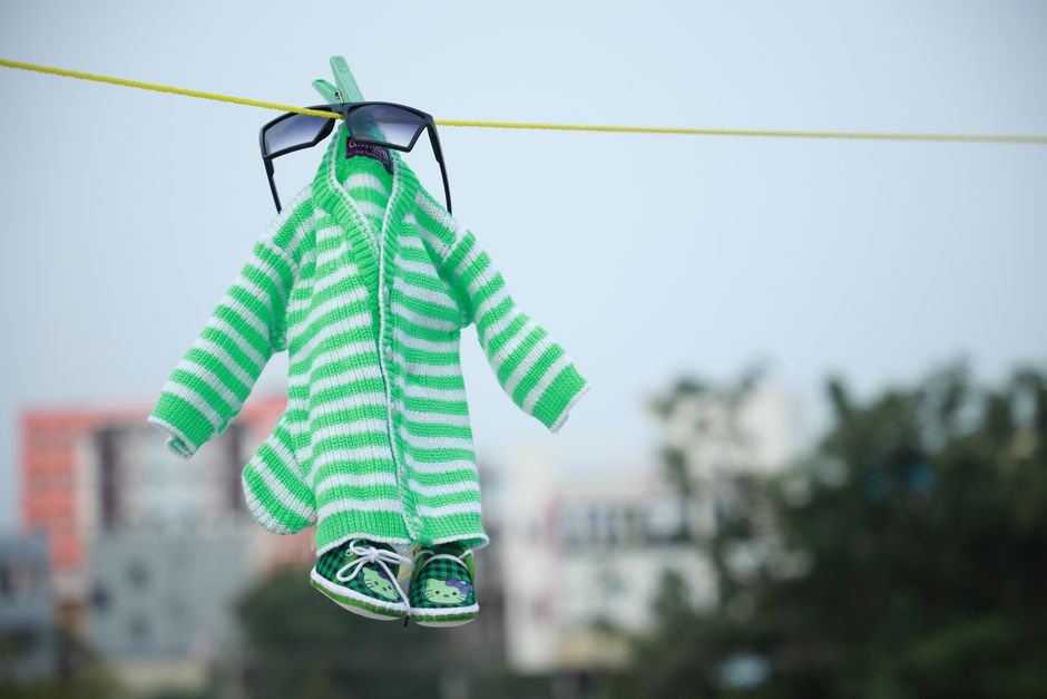 Roupa de bebê pendurada em uma corda de lavanderia, com acessório de óculos escuros e tênis, em um ambiente ao ar livre.