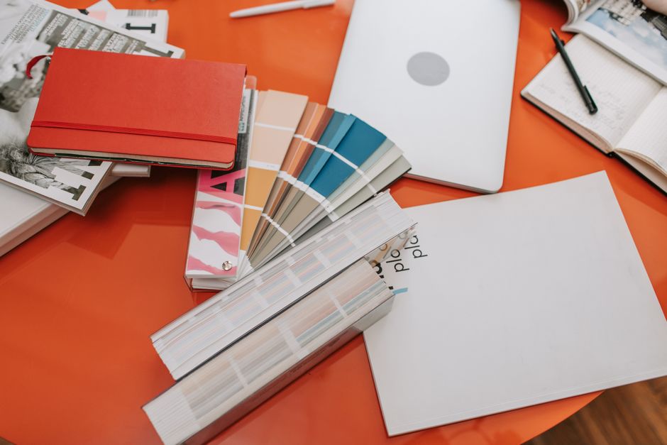 Mesa de trabalho com materiais de design e planejamento, incluindo moldes, catálogos de cores, laptop, e caderno, sugerindo a criação de projetos visuais ou de interiores.
