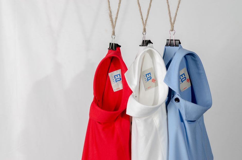Três jalecos de diferentes cores pendurados em cabides de gancho de macramê, representando uniformes profissionais em um ambiente de trabalho de saúde ou laboratorial.