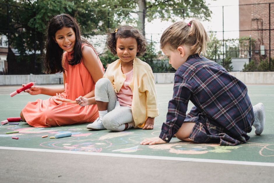 Três crianças brincando juntas ao ar livre, desenhando com giz no chão de uma quadra esportiva.
